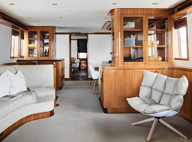 A bright, refined interior of a compact luxury motor yacht or boat, featuring extensive warm timber built-in cabinetry and panelling, white upholstered lounge seating, a sculptural lounge chair, grey carpet, large windows, and a compact galley toward the rear. The space is orderly and highly fitted-out, with mirrored and glazed joinery displaying shelving and storage.