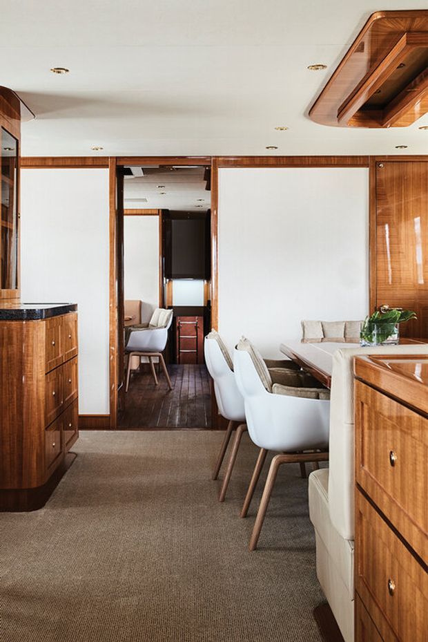 A warm, refined dining and living interior with extensive glossy timber joinery, white walls and ceiling, recessed downlights, and a carpeted floor. A dining table with white upholstered chairs occupies the right side, while a mirrored doorway opens toward a compact adjoining seating area. Built-in timber cabinetry and a sculptural timber ceiling feature frame the space.