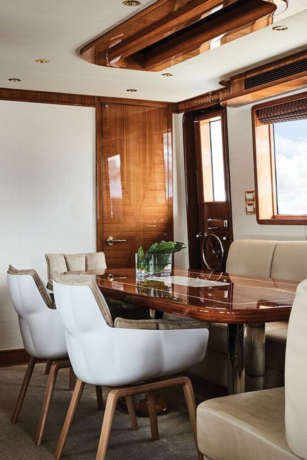 A refined dining and lounge area inside a luxury vessel, featuring a glossy dark timber dining table, upholstered dining chairs, built-in cream seating, rich timber wall and door panelling, a white ceiling with recessed spotlights, and large windows with blinds. The polished timber ceiling feature and nautical interior detailing create an elegant, yacht-like atmosphere.