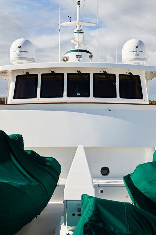 Close-up exterior view of the forward superstructure and wheelhouse of a large white vessel, with dark windows, radar domes, antennas, and covered equipment on the foredeck. The image is dominated by the boat’s exterior rather than an interior room.