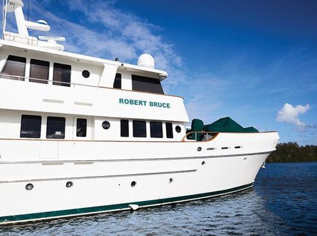 A close exterior view of the large white motor yacht named “Robert Bruce,” showing its multi-level superstructure, dark windows, railings, radar equipment, and green canvas-covered deck area on calm blue water beneath a partly cloudy sky.