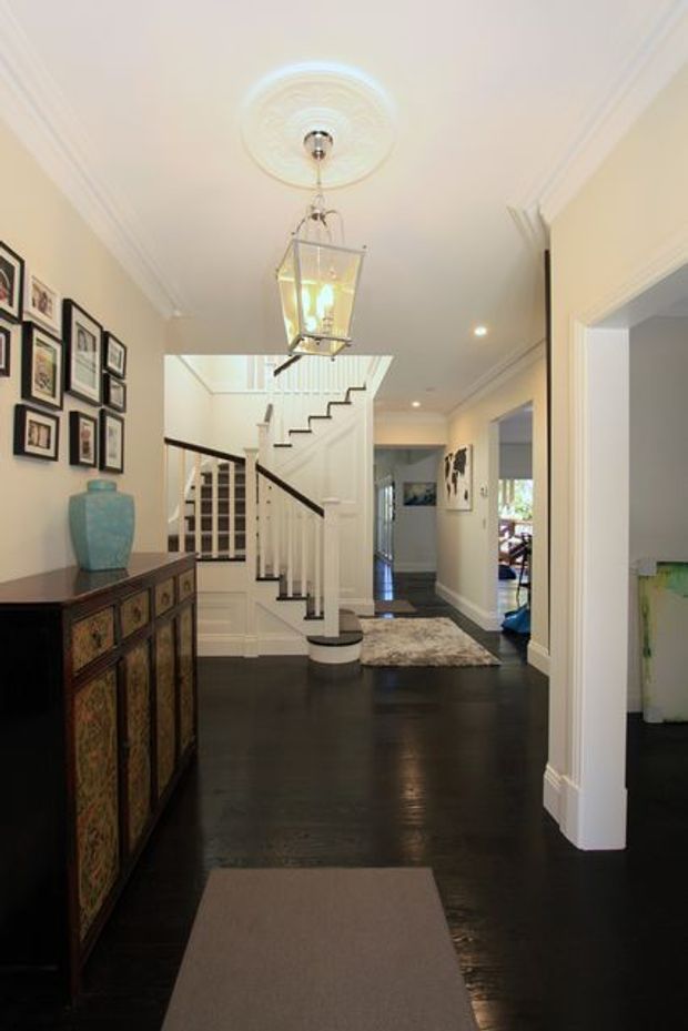 A bright, traditional-style residential entry hall with glossy dark timber flooring, white walls and ornate cornices. A central staircase with white balustrades and dark handrails leads upward beside a skylit landing. Framed artwork forms a gallery wall on the left above a decorative dark timber console, while a lantern pendant hangs from a detailed ceiling medallion. Openings lead toward additional rooms in the background.