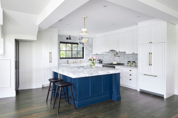 Bright, spacious white kitchen with a large navy-blue island, white shaker cabinetry, marble-look benchtops, grey stone backsplash, dark timber flooring, exposed ceiling beams, and a black-framed window. Three stools sit at the island, with pendant lighting above.