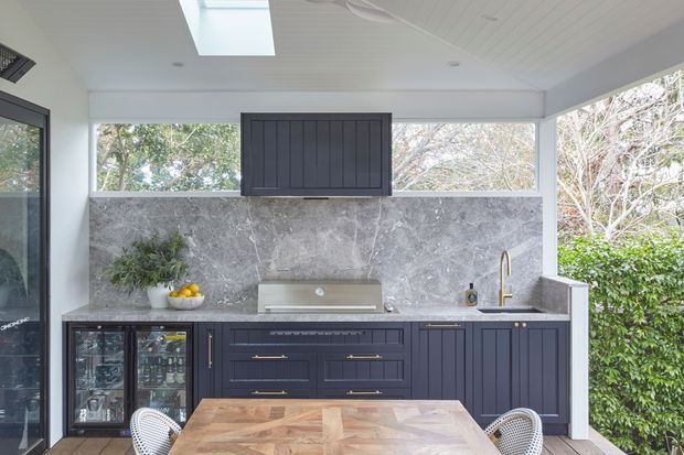 Bright contemporary kitchen with dark blue-grey Shaker-style cabinetry, a large grey marble-look splashback, brass hardware and tapware, integrated rangehood, wine fridge, and timber dining table. The space has generous glazing, a white panelled ceiling, and a skylight overlooking greenery.