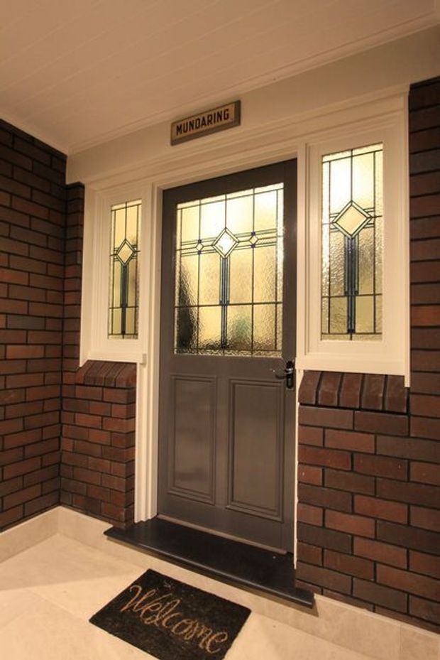 A covered residential entryway featuring a dark painted front door with decorative leadlight glass panels and matching sidelights, set within cream trim and red-brown brick walls. A small address plaque, black threshold, pale tiled floor, and welcome mat complete the traditional entrance.
