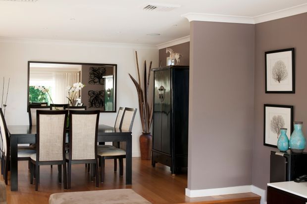 A refined dining area with a dark timber dining table and chairs, set against warm taupe walls and white trim. A large mirror reflects a bright adjoining space, while a tall dark cabinet, decorative branches, framed botanical artwork, and turquoise ceramic vases add visual interest. Timber flooring and a neutral rug create a warm, cohesive interior.