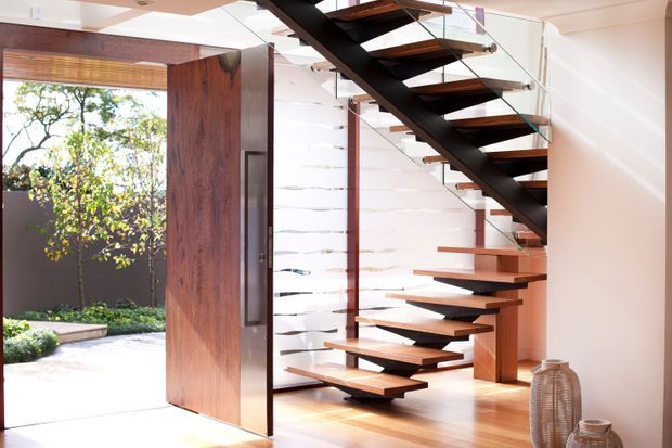 Bright contemporary entryway featuring a sculptural open-riser staircase with dark timber treads, a black structural spine, and clear glass balustrades. A large timber pivot door opens to a landscaped courtyard, while white horizontal wall cladding and timber flooring create a warm, refined interior.