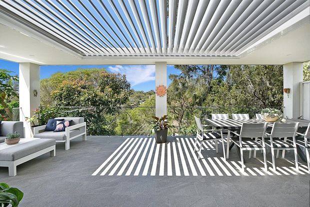 A bright, open-air covered terrace with a motorised louvred roof, grey tiled flooring, white structural columns and glass balustrade. White outdoor lounge seating and a dining table occupy the terrace, with strong striped sunlight and shadow patterns cast across the floor and furniture. Lush trees and blue sky are visible beyond the railing.