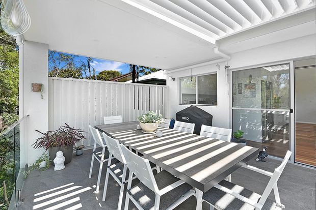 Bright covered outdoor dining area with a large rectangular table and white chairs, set beside glazed sliding doors and a white privacy fence. A louvred pergola roof casts strong linear shadows across the tiled floor and furniture, with potted plants adding greenery.