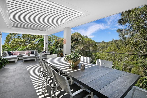 Bright, open-air covered terrace with a large dining table and chairs, an outdoor lounge, grey tiled flooring, white structural columns, and a slatted adjustable roof casting strong linear shadows. The terrace overlooks leafy vegetation and a wooded landscape.