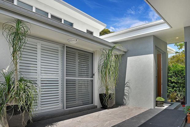 Contemporary residential courtyard or entry area with white rendered walls, flat rooflines, extensive louvred windows and doors, and a recessed timber entry door. Tall potted plants frame the paved approach, with a covered outdoor walkway and dark linear matting creating contrast.