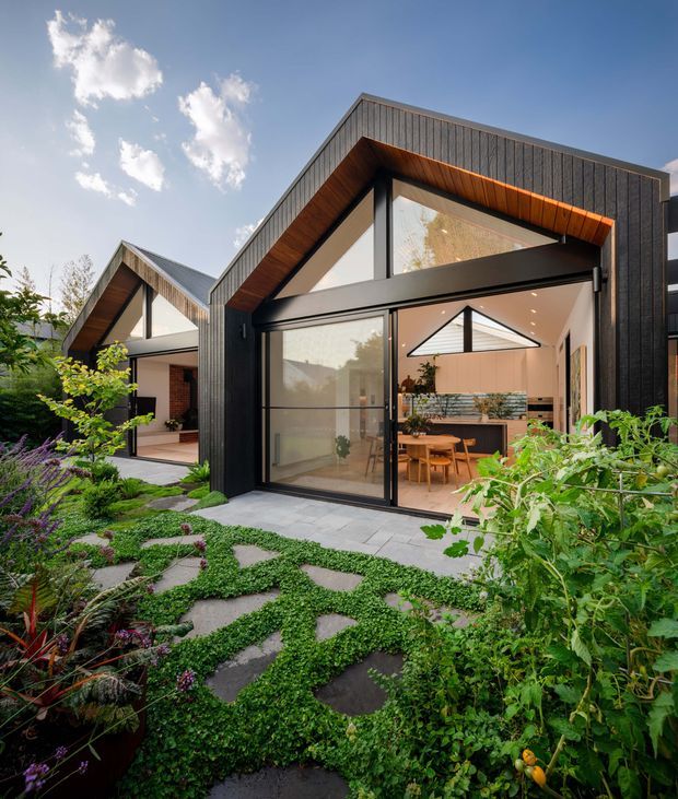 Contemporary garden-facing home exterior with two steeply pitched gabled volumes, dark vertical cladding, black-framed full-height glazing, and warm timber-lined eaves. The open glazed frontage reveals a bright dining and kitchen area, while a landscaped garden with irregular stone stepping paths and dense planting fills the foreground.