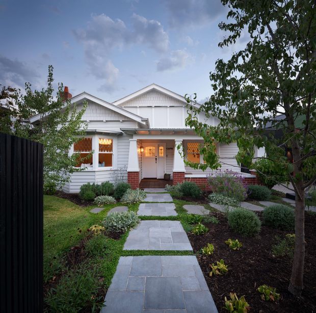 Front exterior view of a charming white weatherboard house with a prominent gabled roof, decorative gable detailing, red-brick base accents and chimney, and warm light glowing from the entry and windows. A landscaped front garden with clipped shrubs, young trees, lawn, mulched planting beds, and a straight grey-paver path leads to the entrance. The home has a traditional cottage or bungalow character.