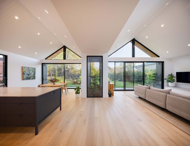 Bright open-plan contemporary living space combining a kitchen, dining area and living room. The room features pale timber flooring, a dark kitchen island, extensive black-framed glazing, triangular clerestory windows, a pitched ceiling with recessed lighting, a dining table, and a large sectional sofa.