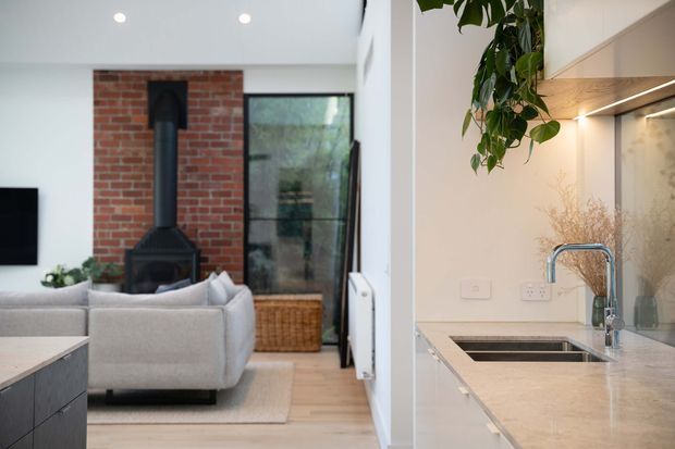 Open-plan interior showing a contemporary kitchen in the foreground and a living room beyond. The kitchen has pale cabinetry, a light stone benchtop, stainless-steel sink and tap, and a subtle backsplash. The living area includes a light sectional sofa, a wall-mounted television, pale timber flooring, and a black wood-burning stove set against an exposed red-brick feature wall. Large glazing brings in greenery and natural light.