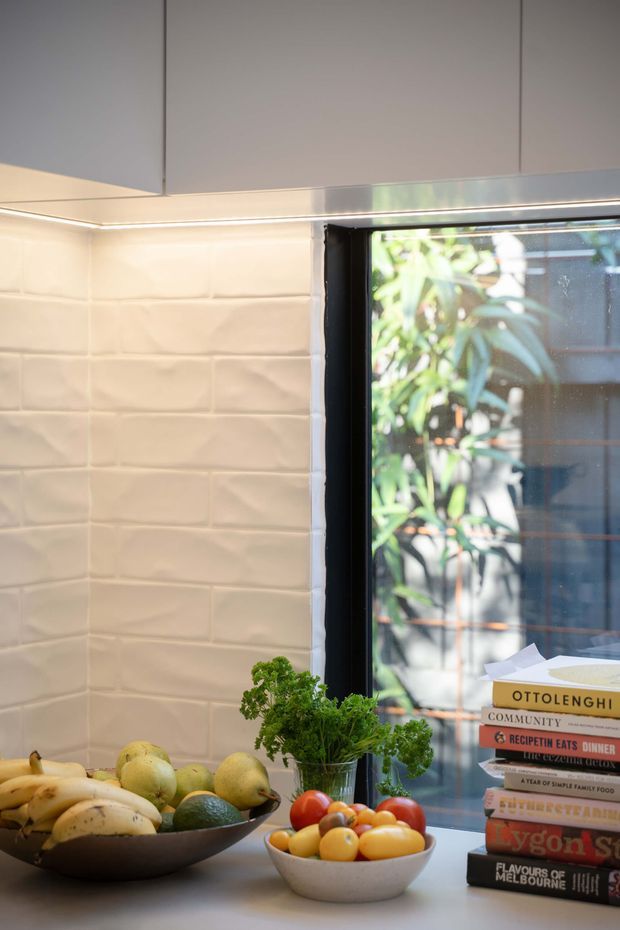 Close-up view of a contemporary kitchen corner with matte grey overhead cabinetry, warm under-cabinet lighting, a white textured subway-tile splashback, and a dark-framed window overlooking greenery. A stone or composite benchtop holds bowls of fruit, herbs, and a stack of cookbooks.