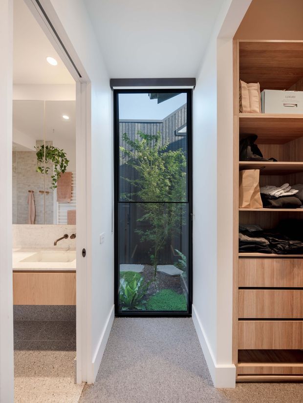 A narrow, bright interior passage with a full-height black-framed window overlooking a small planted courtyard. The hallway has light carpet, white walls and ceiling, and a built-in timber wardrobe with open shelving and drawers on the right. Through the open doorway on the left, a bathroom is visible with a timber vanity, pale stone or tile surfaces, and a walk-in shower.
