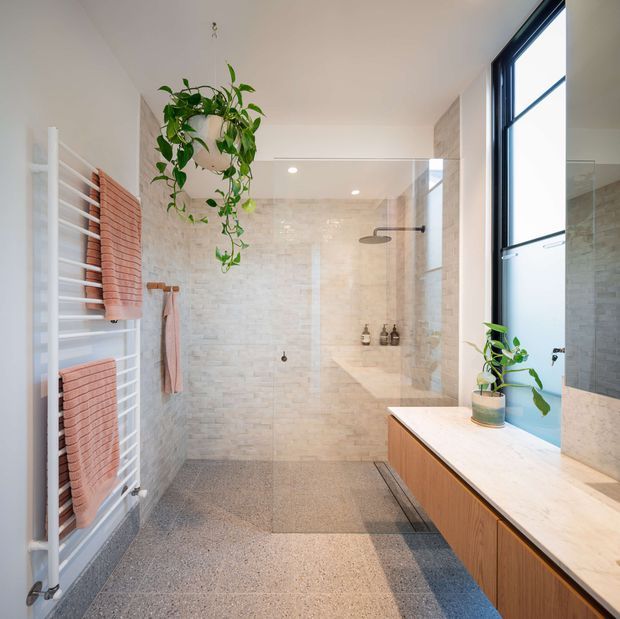 Bright contemporary bathroom with a walk-in glass shower, pale textured wall tiles, terrazzo-style flooring, a timber-veneered vanity with a light stone benchtop, black-framed window, heated towel rail, and hanging greenery. The room is naturally lit and has a clean, refined feel.