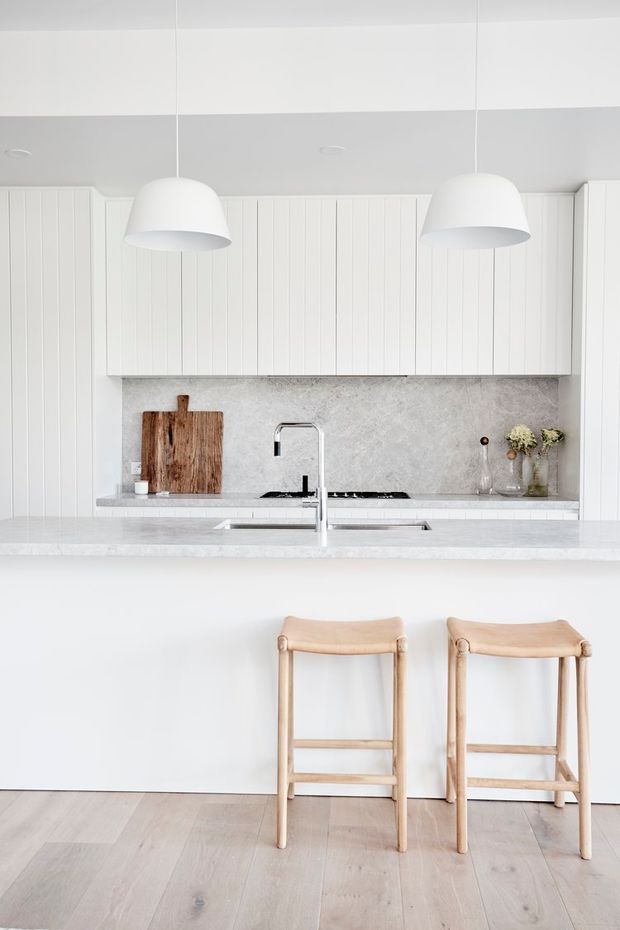 Bright, minimalist white kitchen with a large island and pale timber bar stools. The kitchen features vertically panelled white cabinetry, a pale stone backsplash and benchtop, a chrome tap, and two oversized white pendant lights. Light timber flooring and restrained styling create an airy, calm appearance.