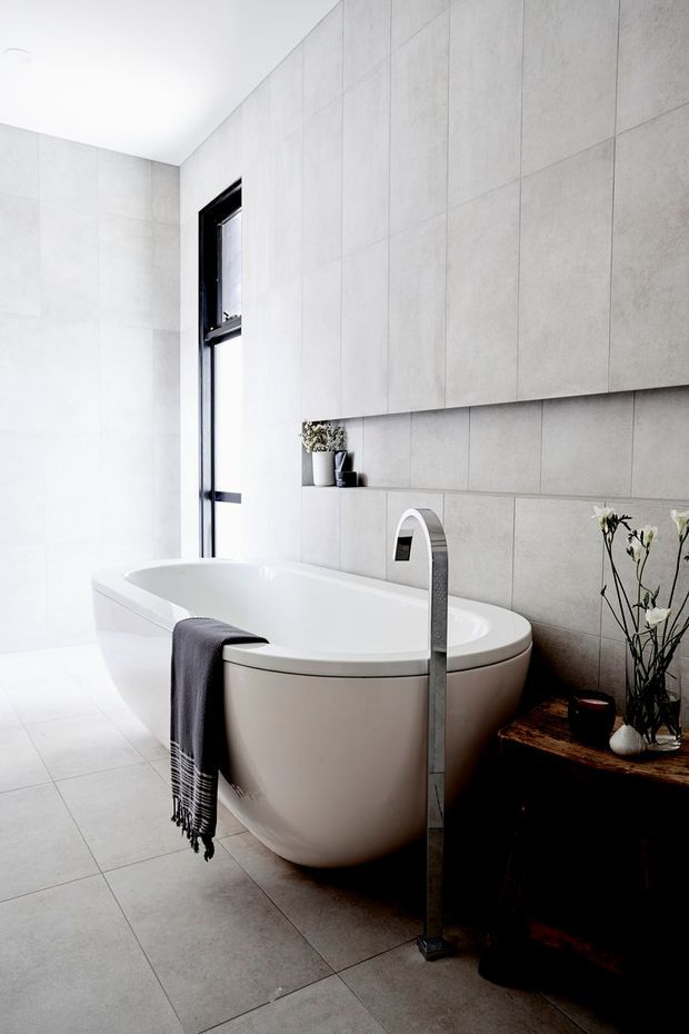 A bright, contemporary bathroom featuring a freestanding oval bathtub, large-format pale grey tiled walls and floor, and a tall brushed metal floor-mounted bath filler. A narrow black-framed window provides natural light, while a dark timber vanity or bench with simple floral styling adds contrast.