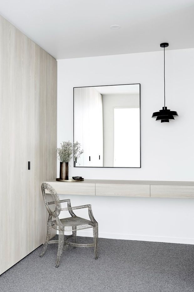 A minimalist, bright interior with full-height pale timber wall panelling and concealed cabinetry on the left, a long floating pale timber console, and a large black-framed rectangular mirror above it. A sculptural black pendant hangs from the white ceiling, while a transparent wire-style armchair and simple decorative vase occupy the foreground. The floor is covered in grey carpet, creating a clean, restrained entry or hallway setting.