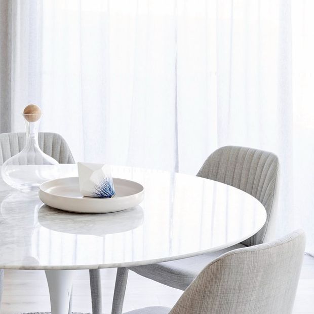 Bright, minimalist dining area featuring a round white marble-look table, upholstered grey dining chairs, sheer floor-to-ceiling curtains, and simple neutral table decor. The space has an airy, refined contemporary aesthetic with abundant diffused natural light.