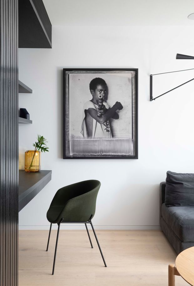 A refined contemporary living room corner with pale timber flooring, white walls, and dark built-in vertical shelving and cabinetry along the left side. A dark green upholstered armchair sits beneath a framed black-and-white artwork, with part of a charcoal sofa and a light timber coffee table visible at right. Minimal black wall lighting and restrained furnishings create a clean, monochromatic interior.