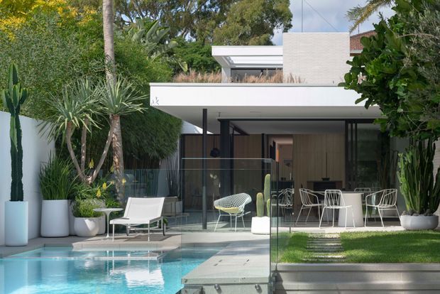 A contemporary flat-roofed house viewed from the landscaped outdoor area, with a glass-walled covered patio opening to a dining and living space. The foreground features a turquoise swimming pool, frameless glass pool fencing, pale paving, lawn, dense tropical planting, and sculptural outdoor furniture. White rendered and brickwork volumes create a clean, modern facade.