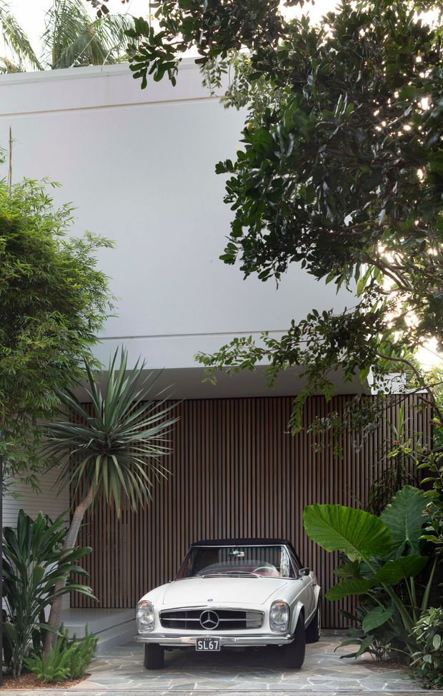 A lush landscaped driveway or entry court leads to a modern residence. A classic white Mercedes convertible is parked in front of a tall vertical timber-slat garage or entry screen, beneath a clean white cubic facade. Dense tropical planting, including palms, yucca-like foliage and broad-leaf plants, frames the approach. The paving is composed of irregular grey stone slabs.