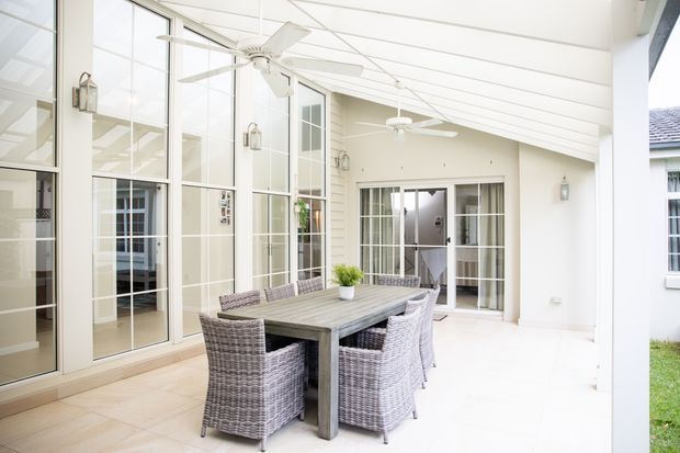 Bright enclosed outdoor entertaining area with a large timber dining table and woven wicker dining chairs. The space has pale tiled flooring, white weatherboard-style walls, extensive multi-pane glazing, a pitched translucent roof, ceiling fans, and lantern-style wall lights. It opens toward a lawn and appears connected to the adjoining house.