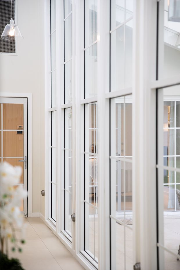 Bright, narrow interior corridor or entry area with a bank of tall white-framed glazed doors or windows featuring multiple panes and slim black lines. A simple timber entry door is visible at the left, with pale tiled flooring, white walls, and a pendant light. The glazing reflects and reveals additional bright interior space beyond, creating an airy, contemporary atmosphere.