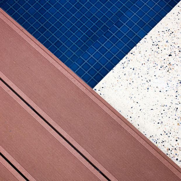 Abstract close-up of an outdoor pool edge featuring deep blue square mosaic tiles, a white terrazzo or aggregate paving surface with multicoloured chips, and reddish-brown ribbed composite decking boards with dark expansion gaps. The image emphasizes contrasting hardscape materials and geometric diagonal composition.