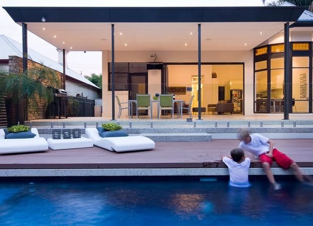 A contemporary single-level pavilion-style home opens onto a broad timber pool deck. The image highlights a covered outdoor entertaining area with a flat roof, slender dark structural posts, full-height glazing, an outdoor dining setting, and a warm-lit interior kitchen or living space beyond. White pool loungers and a deep blue swimming pool occupy the foreground, while adjacent residential fencing and planting frame the scene.