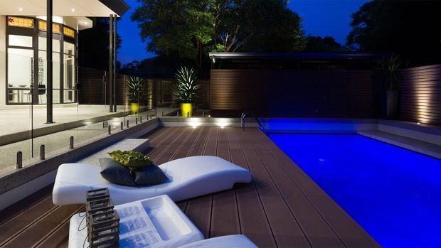 Night-time view of a sophisticated outdoor pool area adjoining a modern glass-walled residence. A vivid blue rectangular pool is surrounded by a broad timber-toned deck, integrated step lighting, dark horizontal wall cladding, sculptural white outdoor lounge seating, and potted plants. The home’s illuminated glazing and clean angular roof forms are visible on the left.