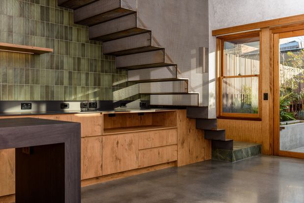 Compact contemporary kitchen and stair area featuring warm timber cabinetry, a dark stone or concrete island, green glazed-tile backsplash, polished concrete flooring, and an open-riser floating staircase with dark timber treads and fine metal balustrade. Timber-framed glazing and a glazed door open toward a planted exterior courtyard.