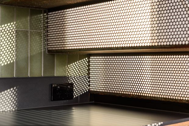 Close-up of a contemporary kitchen work area featuring dark cabinetry, a dark benchtop, and a distinctive perforated metal splashback or screened cabinetry detail. Strong patterned sunlight casts circular shadows across the surfaces, creating a high-contrast industrial atmosphere.