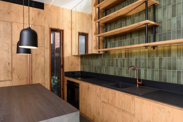 Contemporary kitchen with warm timber cabinetry and wall panelling, a dark benchtop and island, green vertical-format tiled splashback, open timber shelving with black metal supports, and a copper-toned gooseneck tap. A black pendant light hangs over the island, and a glazed internal door adds natural light.