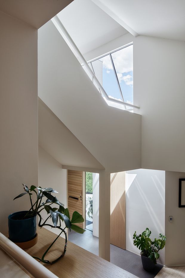A bright, minimalist interior view of a stairway and entry hall. White angular walls and a sculptural stair enclosure frame a large roof skylight, bringing in daylight and views of blue sky. A timber entry door and warm timber stair or landing surfaces contrast with the white walls, while potted plants add greenery. The space has a clean, contemporary architectural character with strong geometric forms and abundant natural light.
