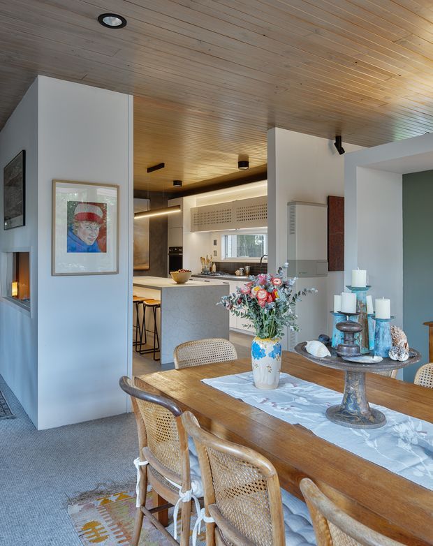 Open-plan dining and kitchen interior with a timber-lined ceiling, warm timber dining table, woven cane dining chairs, and a sculptural stone kitchen island in the background. White walls and built-in joinery are accented by a muted green feature wall, artwork, decorative candles, and a floral arrangement. Recessed and concealed lighting create a warm, inviting atmosphere.