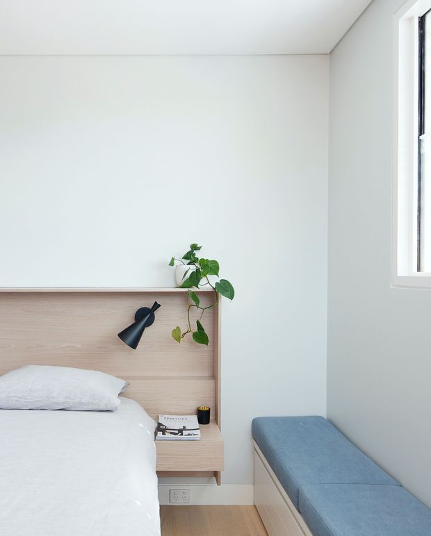 Bright, compact contemporary bedroom corner with a neatly made white bed, pale timber integrated headboard and bedside shelf, black wall-mounted reading light, trailing potted plant, and a long blue upholstered bench beneath a tall window. The room has white walls and ceiling, light timber flooring, minimal furnishings, and abundant natural light.