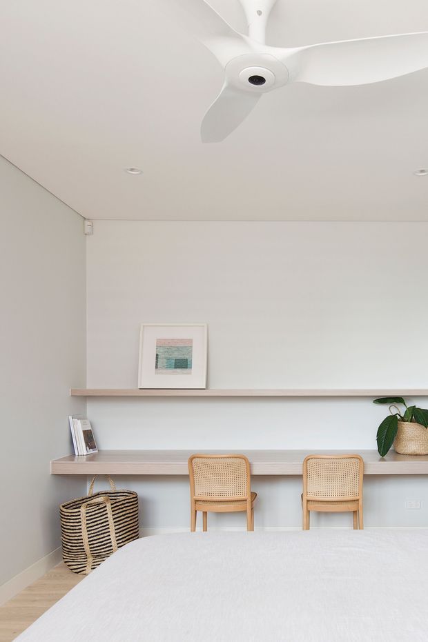 A bright, minimalist bedroom workspace with a pale built-in desk and long floating shelf spanning the wall. Two woven cane-back chairs sit at the desk, with a small framed artwork, books, and a potted plant adding understated detail. A large white bed occupies the foreground beneath a white ceiling fan, and the room is finished in soft neutral tones with recessed ceiling lights and light timber flooring.