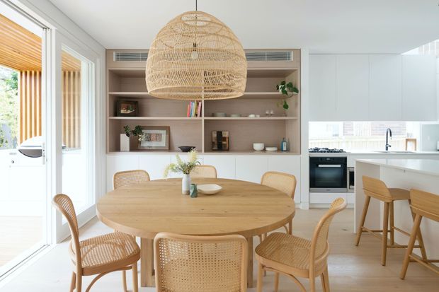 Bright open-plan dining area adjoining a contemporary white kitchen. A round light-wood dining table is surrounded by woven cane chairs, with a large woven pendant light overhead. Built-in timber shelving and cabinetry line the wall behind the table, while full-height glazing opens toward an outdoor area with vertical timber screening. The palette is light, warm, and natural.