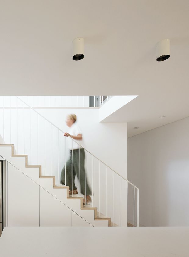 Bright minimalist interior view of a modern stairway. A white-painted staircase with pale timber stair treads and slender white metal balustrades rises through a light-filled open void. The surrounding walls and ceiling are white, with simple recessed and cylindrical ceiling lights. A blurred person is walking on the stairs, emphasizing the vertical circulation space and clean architectural lines.