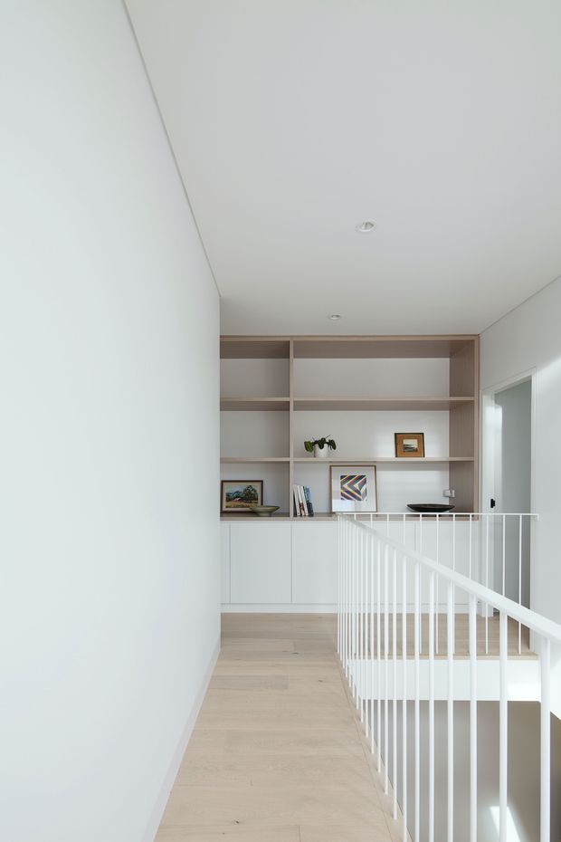 A bright minimalist upper-floor landing or hallway with pale timber flooring, white walls and ceiling, recessed downlights, and a white metal balustrade overlooking the stairwell. A full-height built-in shelving and cabinetry unit in a light timber finish occupies the far wall, displaying a few books, framed artwork, and small decor. An open doorway is visible to the right.