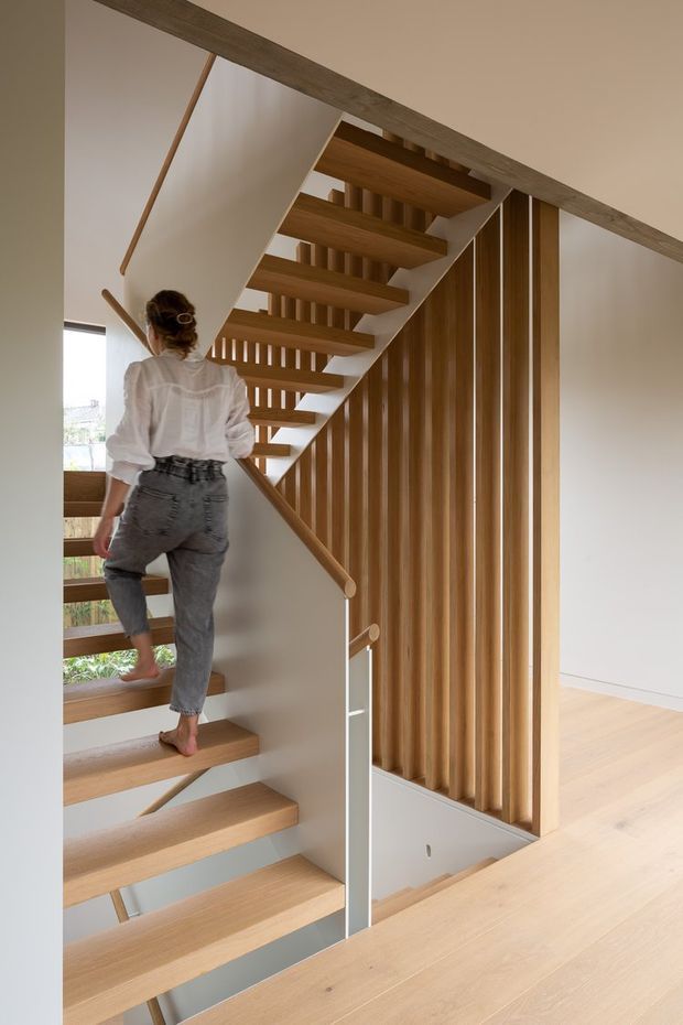 Interior view of a contemporary stairway with open-riser light timber treads, slim white stringers, timber handrails, and a vertical timber-slat balustrade or screening wall. The stair rises through a bright, minimalist upper-level space with pale timber flooring and white walls. A person is shown ascending the stairs.