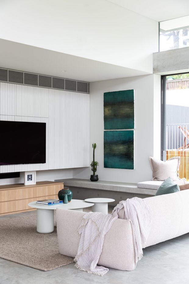 Bright contemporary living room with a high ceiling and large glazed opening. The room features a white vertically ribbed TV feature wall with integrated ventilation, a timber media console, concrete-look window seat, pale upholstered sofa, round white coffee tables, textured rug, and abstract teal artwork. Natural light creates an airy, calm interior.