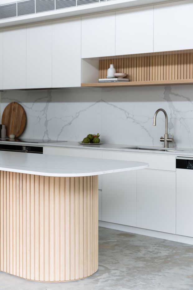 A bright contemporary kitchen featuring white flat-panel cabinetry, a large white stone island with a rounded end, and vertical light timber battens on the island front and open shelf backing. The rear wall has a white marble-look splashback with grey veining, a stainless steel gooseneck tap, integrated appliances, and a grey concrete-look floor. A linear air-conditioning vent runs above the cabinetry.