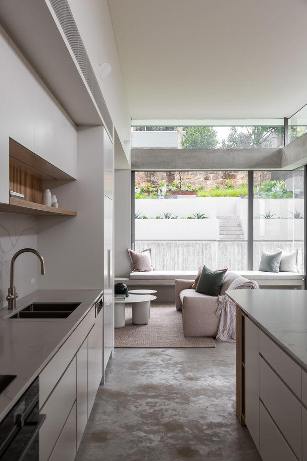 A contemporary open-plan kitchen and living area viewed from the kitchen. The kitchen has sleek pale built-in cabinetry, dark stone benchtops, an integrated cooktop and sink, and open timber shelving. A large island occupies the foreground, while the living area beyond features a neutral sofa, upholstered chair, small round tables and a textured rug. Full-height glazing and a high-level window bring in abundant natural light and overlook a landscaped, terraced courtyard with exposed masonry and concrete elements.
