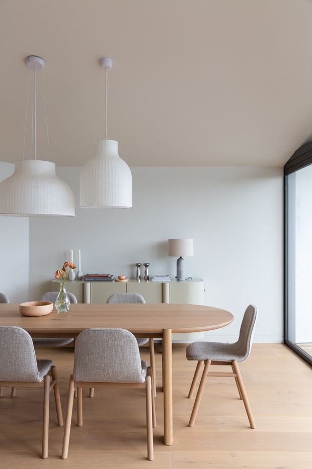 A bright, refined dining room with a large oval timber dining table surrounded by upholstered dining chairs. Two oversized white ribbed pendant lights hang above the table, while a low light-toned sideboard with decorative objects and a table lamp sits against the pale wall. Full-height dark-framed glazing on the right brings in abundant natural light. The room has a calm, minimalist Nordic-inspired aesthetic with warm timber flooring and furniture.