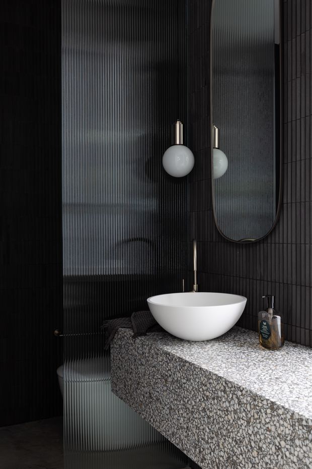 A dark, refined bathroom featuring a floating terrazzo vanity with a white vessel basin, slim wall-mounted tap, black vertically ribbed wall surfaces, and a large rounded-rectangle mirror. A spherical pendant light hangs beside the mirror, while a translucent ribbed glass shower enclosure is visible at left.